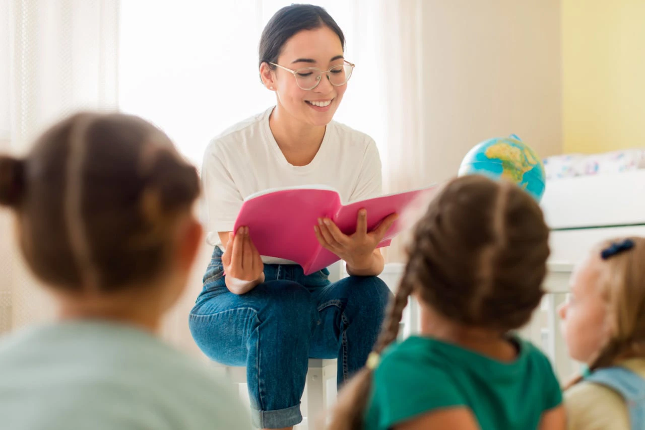 A Woman With Glasses, Sitting On A Stool, Reads A Pink Book To Three Small Children. The Room Is Brightly Lit, With A Globe On A Table Nearby.