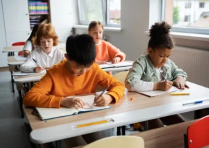 Four Children In A Classroom, Writing In Notebooks. Focused Atmosphere, Natural Light From Windows. Diverse Group, Casual Attire, Learning Environment.